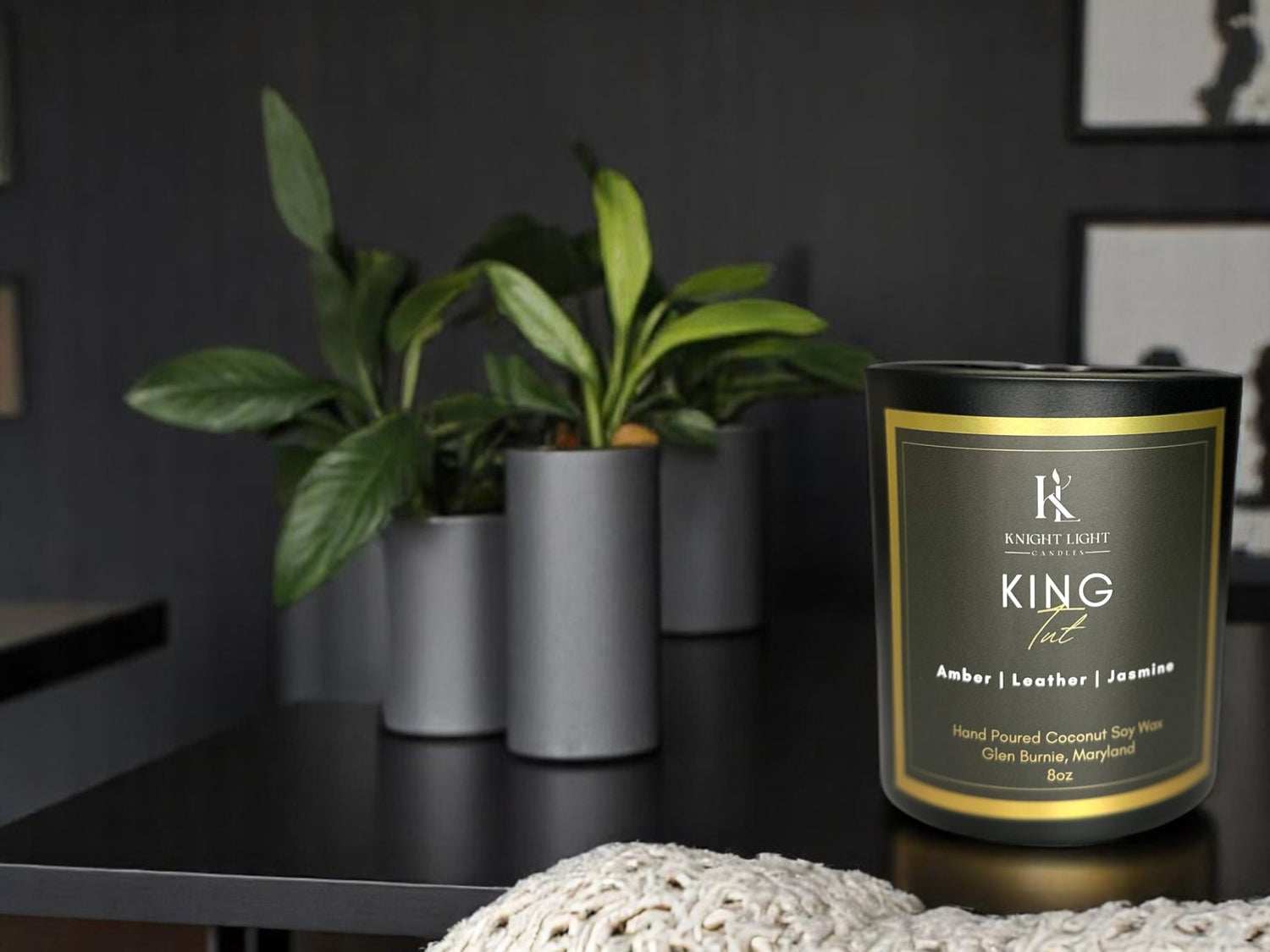 Knight Light Candles King Tut candle on a dark table in a modern home setting with potted plants and cozy knit fabric, highlighting a luxurious and masculine ambiance.